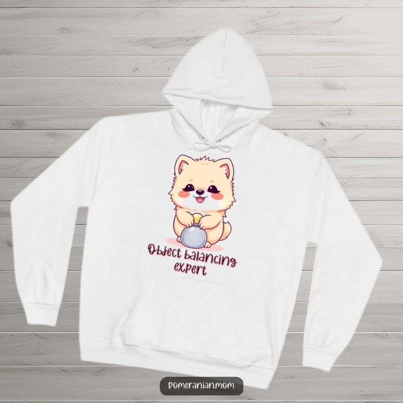 Funny Pomeranian hoodie showing a happy dog balancing a small shiny object, radiating skillful focus and cozy charm.